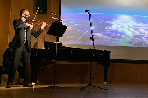 Matthew Lamb in business suit and mask playing the violin in a concert hall